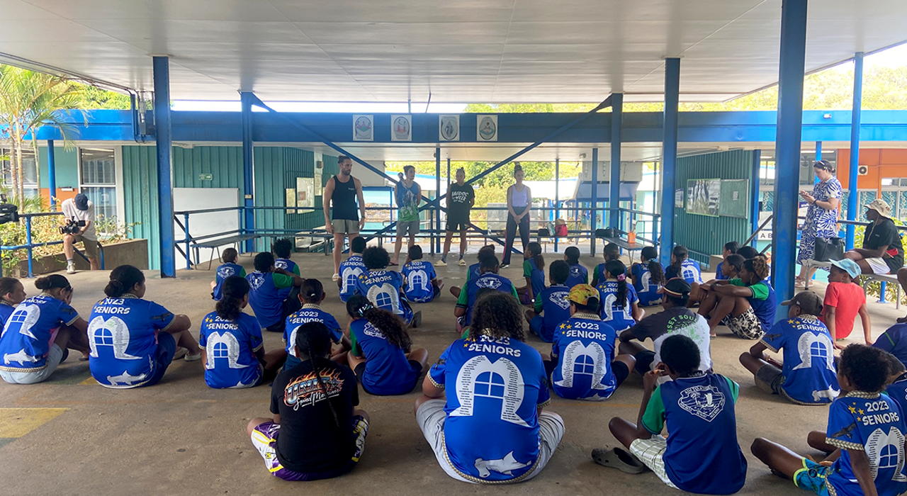 Four dancers stand in front of a large group of year five students in a covered outdoor area. The students sit on the floor facing the dancers. 