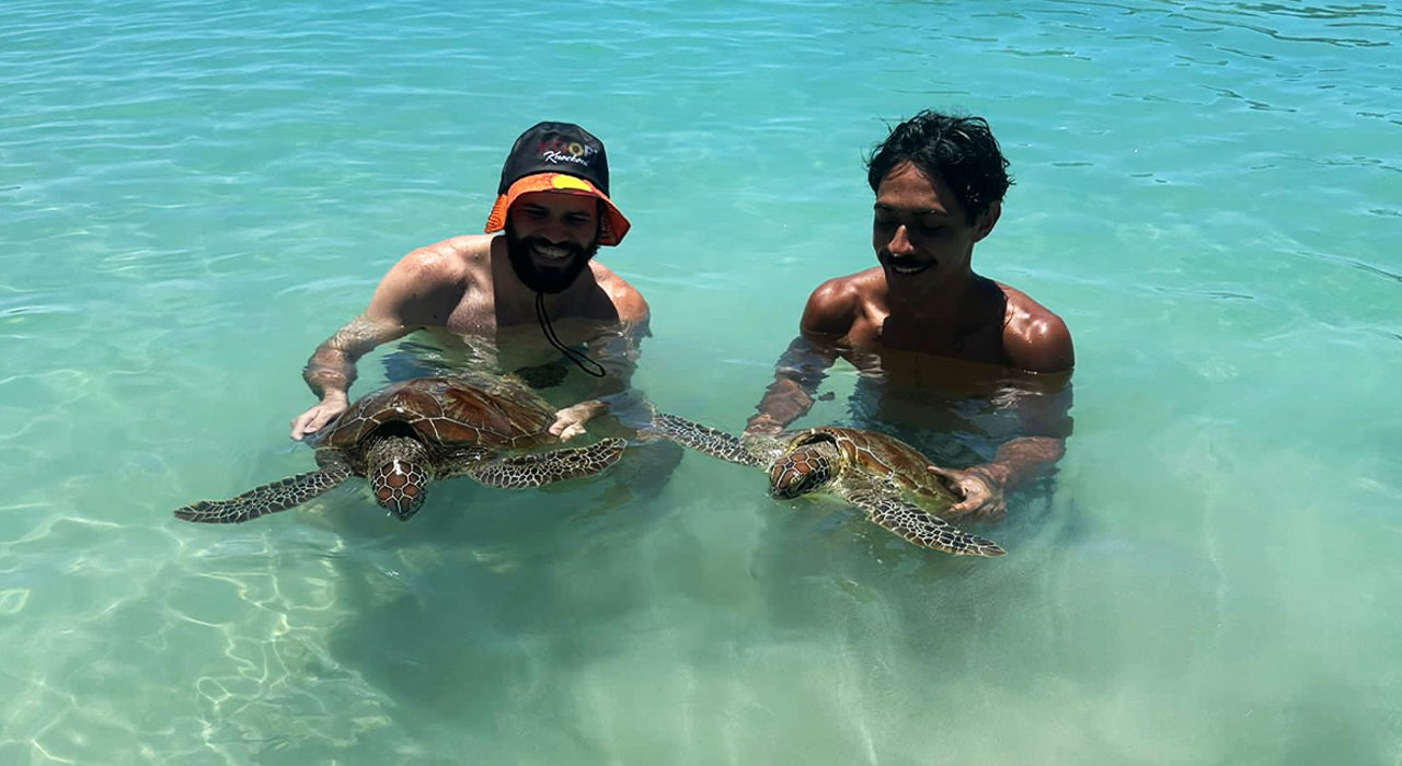 Dancers Jesse Murray and Daniel Mateo in shallow water in the Torres Strait Islands, each smiling, looking down and holding a large green turtle.