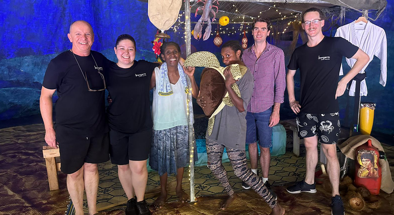 The cast and crew of Waru stand arm in arm, smiling at the camera, standing on the set on stage at the sports complex on Thursday Island.